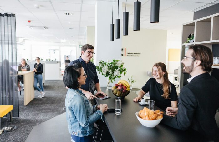 Vier Menschen stehen im öffentlichen Büroraum neben hohen Tisch. AM tisch sind Gebäck, Obstkorb und Blumen. Im Hintergrund ist weitere vier Menschen sichtbar