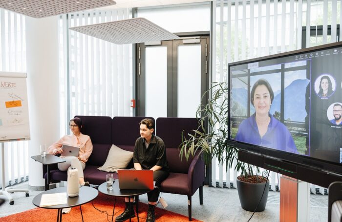 Zwei Menschen sitzen in Konferenzraum mit Laptop und auf Großen Bildschirm läuft Videocall