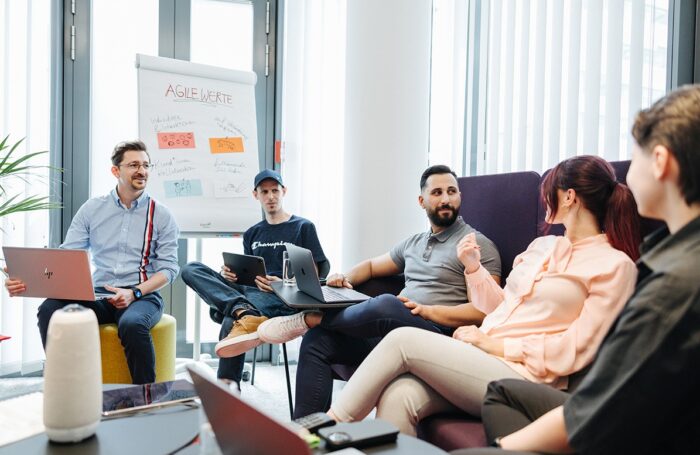 Fünf Menschen sitzen im Büroraum auf Couch und unterhalten. Drei Menschen haben Laptops und eine haltet im Hand iPad