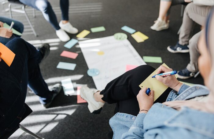 Ein Kreis von Menschen, die in einer Workshopsituation sind und etwas auf farbige Zettel schreiben. Auf dem Boden liegt ein Flipchart-Blatt.