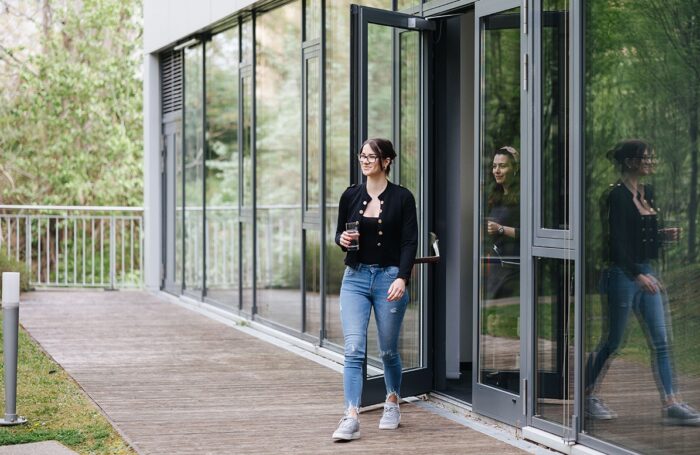 Zwei Frauen verlassen ein Gebäude durch eine Glastüre und gehen in einen Innenhof. Das Gebäude hat eine große Fensterfront.