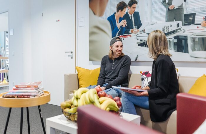 Zwei Personen sitzen auf einem hellen Sofa und unterhalten sich. Davor stehen zwei Tische: einer mit Büchern, einer mit einem großen Korb voll frischem Obst.