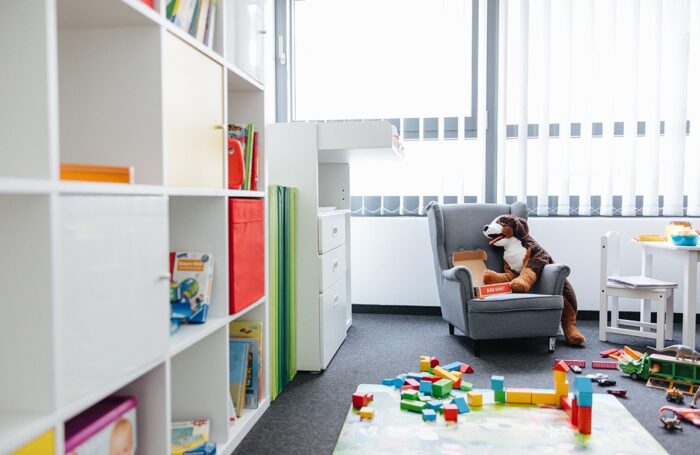 Ein Büro mit Kinderspielzeug: Links steht ein Regal mit Büchern. In der Mitte ein gemütlicher Sessel für Kinder mit einem großen Plüschtier. Rechts steht ein kleiner Tisch mit einem Stuhl, auf dem Boden ist Spielzeug verstreut.