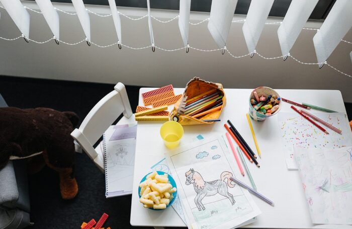 Auf einem Kindertisch liegen Zettel mit ausgemalten Bildern, Buntstifte und Bausteine. Dazwischen stehen ein Becher und eine Schüssel mit Snacks.