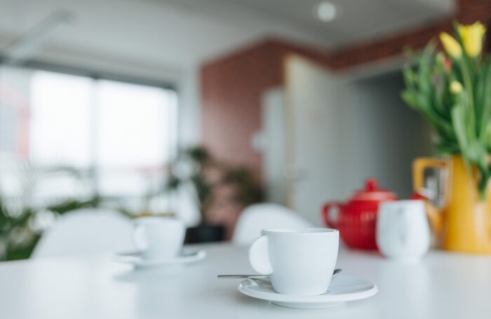 Auf einem Tisch stehen weiße Kaffeetassen, eine rote Teekanne und eine gelbe Blumenvase mit Tulpen.