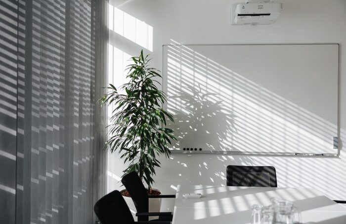 Ein leerer Konferenzraum mit einem großen Tisch, Sesseln, Beamer und Whiteboard. In der Ecke steht eine Pflanze. Durch einen Lamellenvorhang strahlt die Sonne in den Raum.