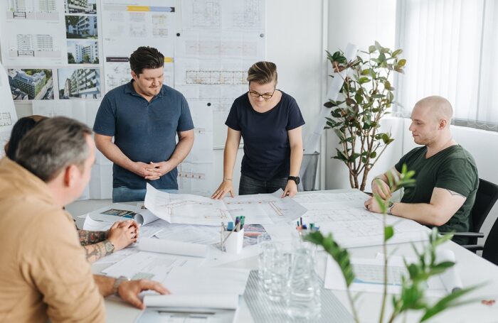 Fünf Personen sitzen und stehen rund um einen Tisch. Auf dem Tisch sind Architekturpläne ausgebreitet. Im Hintergrund hängen Pläne und Bilder an der Wand.