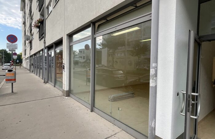 Die Fassade eines Gemeindebaus. Im Erdgeschoss gibt es große Glasfronten. Daneben verläuft eine Straße, an der Autos parken.